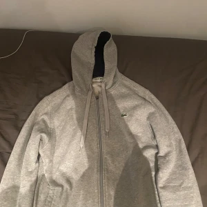Grå zip-hoodie från Lacoste - Grå hoodie med dragkedja från Lacoste. Klassisk krokodillogga på bröstet och justerbar huva med snören. Perfekt för en avslappnad stil och enkel att matcha med det mesta.