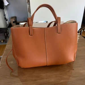 Brun handväska från Polène - Jag köpte den från Plick. Väskan har en stilren design med ljus insida och guldtext på utsidan. Perfekt för dig som vill ha en rymlig och trendig accessoar. Ej äkta