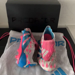 Adidas Predator fotbollsskor i vitt och rosa - Säljer ett par Adidas Predator fotbollsskor i vitt med rosa och blå detaljer. Skorna har en modern design med mönstrade inslag och platt sula med dobbar för bra grepp på planen. Perfekta för dig som vill sticka ut på fotbollsplanen!