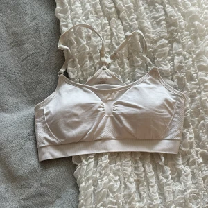 Vit sport-bh från H&M - Säljer en vit sport-bh från H&M med justerbara axelband och ribbade detaljer. Bh:n har en sportig design med bred resår nedtill och lätt rynkning framtill för extra passform. Perfekt för träning eller vardag.