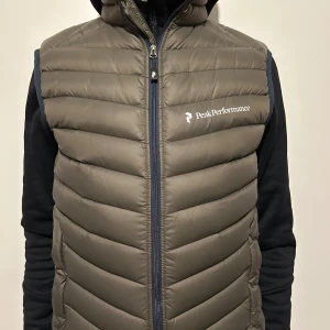 Olivgrön dunväst från Peak Performance - Säljer en olivgrön dunväst från Peak Performance med dragkedja framtill och logga på bröstet. Västen är quiltad och har en sportig look, perfekt för lager-på-lager-stil. Klassisk passform och lätt att matcha med olika outfits.