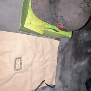 Svart keps från Gucci med monogram - Snygg svart keps från Gucci med klassiskt GG-monogram och grönt/rött band på sidan. Kepsen är i storlek S (57 cm) och har en exklusiv känsla med märkets logga invävd i tyget. Perfekt accessoar för att lyfta din outfit. Den är lite solbränd men det går och fixa och boxen är lite sönder men det spelar inte roll.