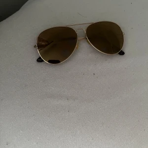 Guldiga pilotbågar solglasögon - Snygga solglasögon med klassisk pilotform och guldiga metallbågar. Glasen är bruna och bågarna har tunna skalmar med mörka ändar. Perfekt accessoar för en cool stil.