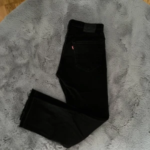 Svarta Levis jeansbyxor W31 L30 - Säljer ett par svarta Levis jeans med klassisk femficksdesign och Levis-logga på bakfickan. Byxorna har normal passform och är tillverkade i jeansmaterial. Uppklippta längst ner så sorleken är ungefär 31/31. Två små hål längst nere där byxorna vart uppklippta (syns inte när man har på sig de). Perfekta för dig som gillar stilrena svarta jeans.