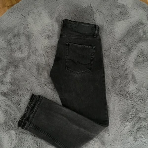 Mörkgråa jeans från Jack & Jones - Säljer ett par mörkgråa jeans från Jack & Jones med klassisk femficksdesign och normal passform. Storleken är 30/30 men de har klippts upp längst ner så storleken är ungefär 30/31. Jeansen har knapp och dragkedja framtill samt bälteshällor. Perfekta till många olika outfits.