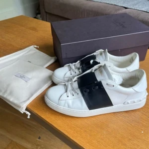 Valentino sneakers med svart detalj - Säljer ett par vita Valentino sneakers med bred svart rem över vristen och ikoniska nitar på hälen. Skorna har rund tå, platt sula och klassisk snörning. Perfekta för dig som gillar stilrena och lyxiga sneakers.