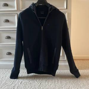 Mörkblå cardigan jacka från Bugatti - Säljer en mörkblå Cardigan jacka från Bugatti med dragkedja framtill och två fickor. Jackan har huva och är tillverkad i ett stickat material med detaljer i syntet. Perfekt för en stilren och avslappnad look. Priset är inte hugget i sten och det är bara o fråga om fler bilder🤩