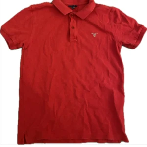 Röd pikétröja från Gant - Säljer en klassisk röd pikétröja från Gant med liten broderad logga på bröstet. Tröjan har krage och knappar framtill, perfekt för en stilren look. Kortärmad och i mjukt bomullsmaterial.