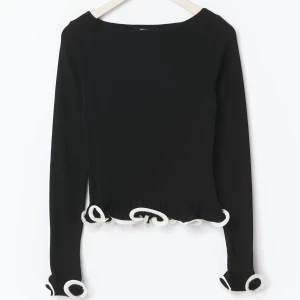 Svart långärmad topp med volangdetaljer - Säljer en svart långärmad topp med vita volangdetaljer vid ärmslut och nederkant. Toppen har en rund halsringning och figurnära passform. Perfekt för dig som vill ha något unikt och stilrent i garderoben.