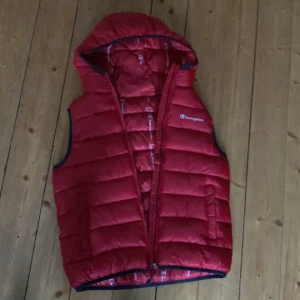 Röd dunväst med huva från Champion - Röd dunväst från Champion med huva och dragkedja. Västen har ett quiltat mönster och logga på bröstet. Insidan är mönstrad med Champion-loggor. Perfekt för lager-på-lager under kyliga dagar användes 1 gång.