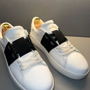 Valentino Garavani Open sneakers i vitt och svart - Snygga vita sneakers från Valentino Garavani med svart band över mitten. Skorna har rund tå, platt sula och klassisk snörning. Sulan är lite trasig på insidan, men ingen som märks. Om man vill skulle jag rekommendera att köpa eget sul inlägg ! Vid funderingar hör av er!