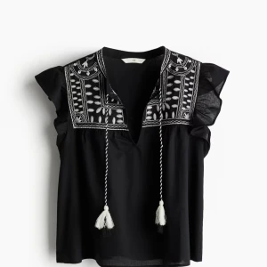 Svart blus med broderi och knytband - Svart blus med vita broderade detaljer upptill och dekorativa knytband med tofsar. Blusen har korta volangärmar och v-ringning. Perfekt för dig som gillar bohemiska detaljer och stilrena färger.