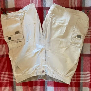 Beiga cargoshorts med fickor - Snygga beiga cargoshorts från Nickelson med flera praktiska fickor på sidorna och baktill. Shortsen har normal passform, elastisk midja och knäppning framtill. Perfekta för varma dagar och avslappnad stil.