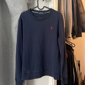 Mörkblå sweatshirt från Polo Ralph Lauren - Säljer en klassisk mörkblå sweatshirt från Polo Ralph Lauren med rund halsringning och röd broderad logga på bröstet. Tröjan har långa ärmar och ribbade muddar vid ärmslut och nederkant. Perfekt för en stilren och avslappnad look.