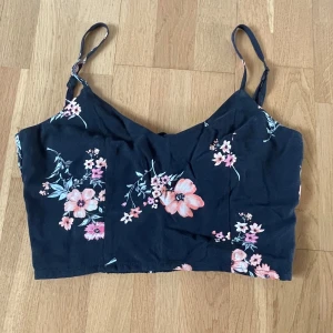 Blommig croppad linnetopp från H&M Divided - Säljer en svart croppad linnetopp från H&M Divided med rosa och vita blommor. Toppen har smala axelband och knytning i ryggen. Perfekt för sommardagar och enkel att matcha med jeans eller kjol.
