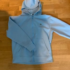 Ljusblå hoodie från Lacoste - Säljer en ljusblå hoodie från Lacoste med dragkedja och klassisk krokodillogga på bröstet. Tröjan har huva och två fickor framtill. Perfekt för en avslappnad och stilren look.