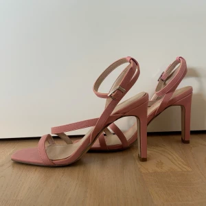 Rosa sandaler med klack och remmar - Säljer ett par stilrena rosa sandaler från Amisu med hög klack och smala remmar över foten och runt vristen. Skorna har fyrkantig tå och justerbart spänne vid ankeln. Perfekta för dig som vill ha en feminin och trendig look.