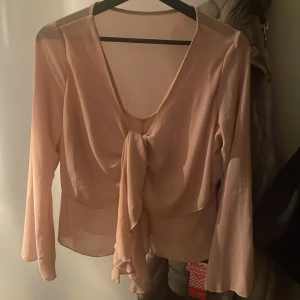 Ljusrosa blus med knyt framtill - Säljer en ljusrosa blus med v-ringning och knyt framtill. Blusen har långa ärmar och är tillverkad i ett tunt, lätt material som ger ett elegant fall.