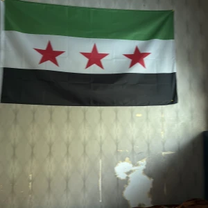 Syriska oppositionens flagga - Säljer en tygflagga med tre horisontella fält i grönt, vitt och svart samt tre röda stjärnor i mitten. Flaggan är rektangulär och passar bra som väggdekoration eller samlarobjekt.