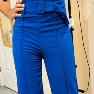 Kornblå vida byxor med bälte - Snygga kornblå byxor med hög midja och matchande bälte med klädd spänne. Byxorna har vida ben och markerade pressveck framtill. Stängs med dragkedja i sidan. Perfekta för en trendig och stilren look.