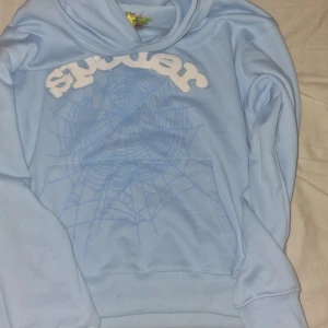 Ljusblå hoddie sp5der - Säljer en ljusblå hoodie från Sp5der med ett stort vitt broderat tryck och spindelnätsmönster på framsidan. Tröjan har huva och magficka, perfekt för en avslappnad stil.
