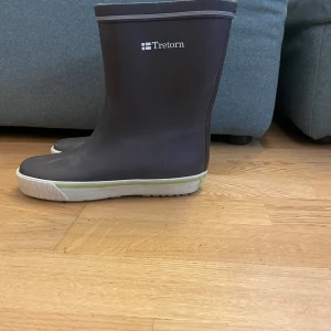 Bruna gummistövlar från Tretorn - Säljer ett par klassiska bruna gummistövlar från Tretorn med vit sula och diskret logga på skaftet. Perfekta för regniga dagar och har en enkel, stilren design med rund tå och platt sula.
