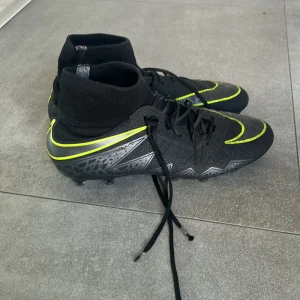 Svarta Nike fotbollsskor med gula detaljer - Säljer ett par svarta Nike fotbollsskor med hög strumpa och gula detaljer. Köpte skorna för ett bra tag sedan precis innan jag slutade i fotboll. Så dom har legat i hyllan nästan helt orörda.