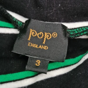 Svart och grönrandig polotröja från Pop England - Säljer en svart polotröja med gröna och vita ränder från Pop England. Tröjan har lång ärm och en klassisk polokrage. Perfekt för dig som gillar stilrena och färgglada plagg med en twist.