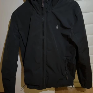 Svart vindjacka från Polo Ralph Lauren - Svart vindjacka med huva från Polo Ralph Lauren. Jackan har dragkedja framtill, två fickor med dragkedja och en liten broderad logga på bröstet. Perfekt för blåsiga dagar. Storlek M men passar S. Priset är inte hugget i sten!!!