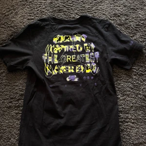 Svart t-shirt från Jordan x Nike med tryck - Säljer en svart t-shirt från Jordan x Nike med stort 'JUMPMAN'-tryck i lila och vitt på framsidan. Baksidan har grafiskt tryck med text och basketdetaljer. Klassisk passform och rund hals. Perfekt för dig som gillar streetwear och basket.
