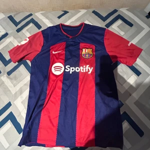 FC Barcelona hemmatröja Nike - Säljer en FC Barcelona hemmatröja med breda blå och röda ränder, Nike-logga och Spotify som sponsor på bröstet. Tröjan har klubbmärket på vänster sida och UNHCR-logga på ryggen. Kortärmad och sportig design.