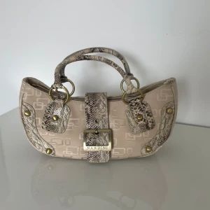 Beige axelväska från Nardini med ormmönstradedetaljer  - Snygg beige axelväska från Nardini med ormmönstrade detaljer och guldfärgade metalldelar. Väskan har dubbla handtag, dragkedja upptill och ett dekorativt spänne framtill. Köpte på loppis för en dag sen för 400kr säljer den då den inte riktigt är min still!!!😁