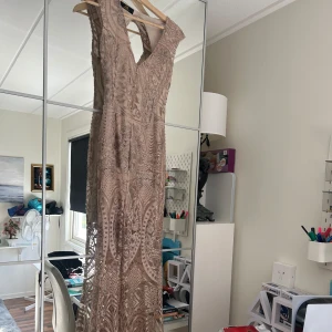 Champagnefärgad långklänning med glittermönster - Säljer en elegant långklänning i champagnefärg med glittrigt, broderat mönster och djup v-ringning. Klänningen har ärmlös design och markerad midja, perfekt för bal eller fest. Materialet känns lyxigt och skimrar snyggt i ljuset.