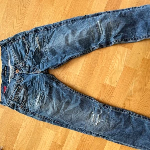 Blå slitna jeans från Guess Jeans - Säljer ett par blå vintage jeans från Guess Jeans med slitningar och detaljerade sömmar. Jeansen har klassisk femficksdesign och normal passform. Tillverkade i 100% bomull för en skön känsla. Perfekta för dig som gillar en avslappnad och trendig stil.