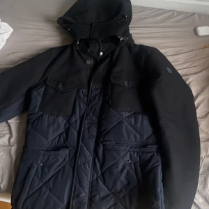 Svart quiltad jacka med huva från GANT - Säljer en svart jacka från GANT med quiltade partier och fyra fickor framtill. Jackan har huva, jackan går för 5000kr i nypris och jag säljer den för endast 1500kr. Den är totalt oanvänd. Anledningen att jag säljer den är för att det var en gåva till mig som inte riktigt är min smak