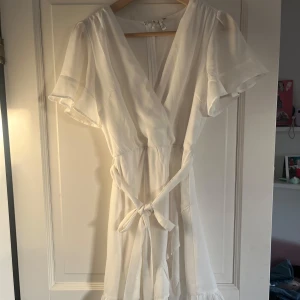 Vit klänning från Nelly🩷 - Säljer en söt vit omlottklänning med volangdetaljer och knytband i midjan. Klänningen har korta, vida ärmar och v-ringning. Perfekt för sommarens alla tillfällen!
