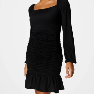 Svart långärmad klänning med rynk och volang från Bubbleroom - Snygg svart klänning från Bubbleroom med lång ärm, fyrkantig ringning och rynkade detaljer framtill. Klänningen har volang nertill och är tillverkad i ett lätt strukturerat material. Perfekt för dig som vill ha en stilren och trendig look. Helt oanvänd med lappen kvar. Skicka gärna prisförslag🩷