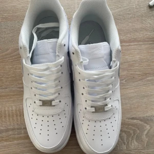 Vita Nike Air Force sneakers - Säljer ett par klassiska vita Nike Air Force sneakers. Skorna har en stilren design med perforerade detaljer på tån och en robust sula. De är utrustade med snörning för en säker passform och har Nike-loggan på hälen.