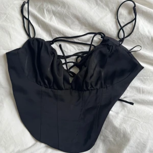 Svart korsettopp med snörning från Gina Tricot - Säljer denna svarta satin topp från Gina Tricot i storlek XS. Den är endast använd vid ett tillfälle därför k väldigt bra skick! Den har snörning i ryggen.  