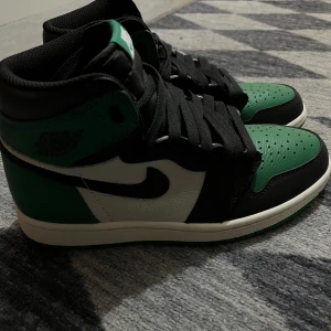 Nike Air Jordan 1 Retro High - Säljer ett par Nike Air Jordan 1 Retro High med svart, grönt och vitt läder. Klassisk hög modell med svart swoosh och svarta skosnören. Perfekta för dig som gillar streetwear och sneakers med attityd. Boxen och allt medföljer. Erbjuder att mötas upp eller att jag fraktar. 
