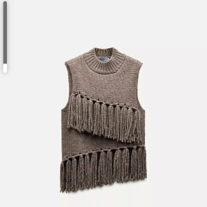 Brun stickad väst med fransar - Säljer en brun stickad väst med grova fransdetaljer i två diagonala rader framtill. Västen har en hög, ribbad krage och är ärmlös. Perfekt för att ge outfiten en unik touch.