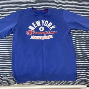Blå sweatshirt från Champion med New York-tryck - Säljer en blå sweatshirt från Champion med stort New York-tryck och texten 'Athletic Department' på bröstet. Tröjan har rund halsringning, långa ärmar och ribbade muddar. Perfekt för en sportig och avslappnad stil.