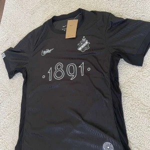 Svart AIK fotbollströja från Nike - Säljer en svart AIK fotbollströja från Nike med trycket '1891' på bröstet och klubbmärket på vänster sida. Tröjan är kortärmad och tillverkad i 100% polyester. Snygga vita detaljer och diskret mönster i tyget.