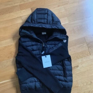 Svart dunväst med huva från Moncler - Snygg svart dunväst från Moncler med stickade partier och quiltad framsida. Västen har huva, dragkedja och Moncler-logga på bröstet. Perfekt för lager-på-lager och stilren look. Äkta. Kvitto finns kontoakta gärna.