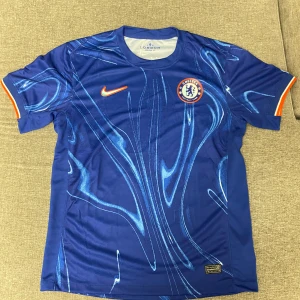 Chelsea FC hemmatröja 2022/23 - Blå Chelsea FC fotbollströja från Nike med abstrakt mönster i ljusblått och vitt. Kortärmad modell med orange detaljer på ärmslut och Nike-logga. Chelsea klubbmärke på bröstet. Tillverkad i lätt och ventilerande material.
