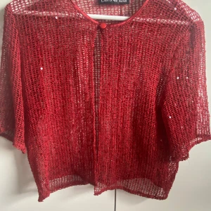 Röd glittrig från Bleu d'Azur - Säljer en röd nätblus från Bleu d'Azur med glittriga paljettdetaljer. Jackan har en enkel knäppning vid halsen och är tillverkad i ett lätt, transparent material. Perfekt för att lägga till lite extra till din outfit.
