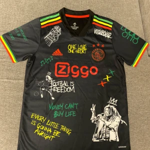Ajax x Bob Marley svart fotbollströja - Säljer en svart Ajax fotbollströja från Adidas med Bob Marley-tema. Tröjan har färgglada ränder på ärmarna, tryck med Bob Marley, Ajax-logga och Ziggo sponsor. Detaljer i rött, gult och grönt samt flera inspirerande texter och motiv.
