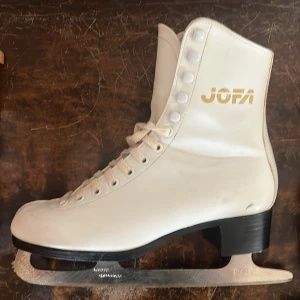 Vita konståkningsskridskor från Jofa - Säljer ett par klassiska vita konståkningsskridskor från Jofa med snörning och svart klack. Skridskorna har rund tå och är tillverkade i syntetmaterial. Perfekta för dig som vill åka skridskor med stil.