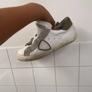 Vita sneakers från Philippe Model - Säljer ett par vita sneakers från Philippe Model med grå mockadetaljer och rund tå. Skorna har platt sula och snörning, samt en ikonisk sköld på sidan. Perfekta för en avslappnad och stilren look.