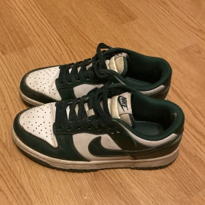 Nike Dunk Low i grönt och vitt - Säljer ett par Nike Dunk Low sneakers i klassisk grön och vit färgkombination. Skorna har lågt skaft, perforerad tå och vit sula. Ikonisk Nike Swoosh på sidorna och snörning framtill. Perfekta för dig som gillar streetwear och sportig stil.